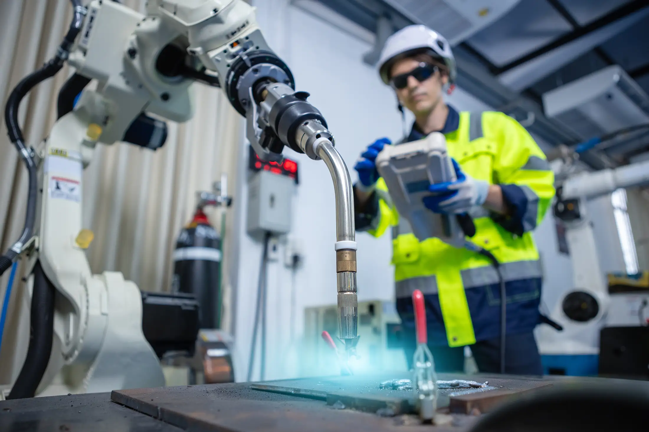 Industrial & Research Pressure Vessel Applications Engineer operating a robotic welding system in an industrial lab, wearing safety gear and controlling the process with a handheld device under bright blue arc light.