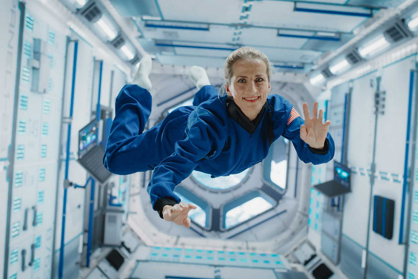 Smiling astronaut in a blue flight suit floating in zero gravity inside a spacecraft module, surrounded by control panels and illuminated equipment.
