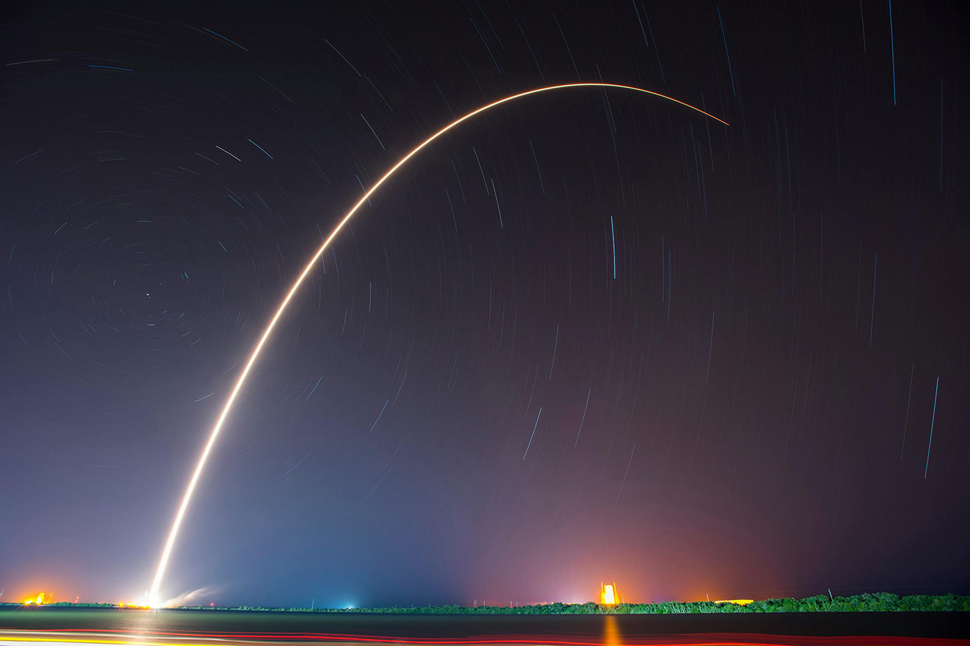 Long-exposure photograph of a rocket launch at night, showing a bright curved arc of light rising into the sky with star trails visible against the dark background.