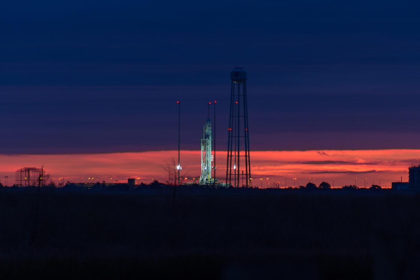 Rocket on a launch pad illuminated at dusk, with a deep blue sky and vivid orange-red sunset on the horizon, surrounded by support towers and lights at a space launch facility.