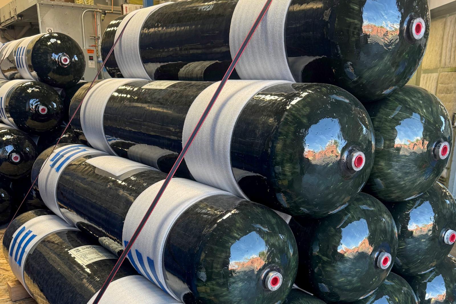 Stacked Type 3 composite CNG cylinders with filament-wound finish and safety labels, secured with straps for transportation.